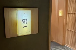 Entrance to Ginza Sushi Ichi. The focus is on a rectangular, backlit frosted glass sign mounted on a light wood panel against a dark, textured wall. The sign features elegant Japanese calligraphy and the English name "Sushi ichi" at the bottom. To the right, a vertical wooden slat door is slightly ajar, revealing a glimpse of a soft, peach-colored interior hallway and another smaller glowing sign, creating a sense of exclusive entry.