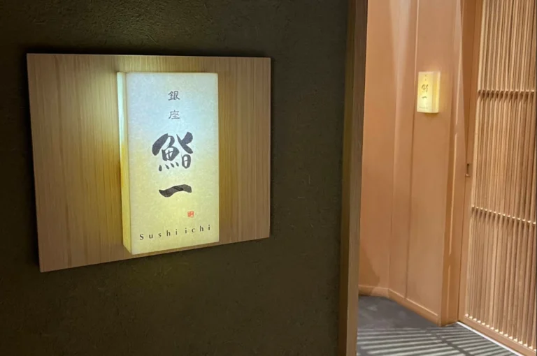 Entrance to Ginza Sushi Ichi. The focus is on a rectangular, backlit frosted glass sign mounted on a light wood panel against a dark, textured wall. The sign features elegant Japanese calligraphy and the English name "Sushi ichi" at the bottom. To the right, a vertical wooden slat door is slightly ajar, revealing a glimpse of a soft, peach-colored interior hallway and another smaller glowing sign, creating a sense of exclusive entry.