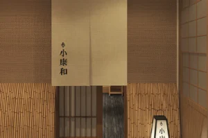 A minimalist Japanese restaurant entrance featuring a light beige fabric noren curtain printed with black kanji characters. The lower walls are clad in vertical bamboo poles, while the upper walls have a textured woven pattern. A warm, glowing floor lantern sits to the right, and a sliding shoji door is visible in the background.