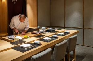 A high-angle, wide shot of an elegant, minimalist omakase sushi counter crafted from light-toned wood. A sushi chef in a crisp white uniform and face mask stands behind the counter, focused on meticulously slicing fresh fish. The counter is impeccably set for several guests with black placemats, white napkins, and wooden chopsticks, while a warm, neutral-toned wall and a textured grey stone pillar create a sophisticated, serene atmosphere in the background.