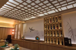 This wide, eye-level indoor shot captures the serene interior of Sushi Kawasemi in Singapore, showcasing a sleek wooden sushi counter beneath a traditional Japanese-style gridded shoji paper ceiling that softly diffuses light. The back wall features a minimalist, misty landscape mural in muted grey tones depicting reeds and a prominent kingfisher bird in mid-flight carrying a twig. To the right, a tall, recessed wooden shelving unit is beautifully illuminated, displaying four rows of various sake bottles with distinct colorful labels and dark glass. Below the mural on the long wooden counter sit a rustic wooden sculpture with a blue glass center and a piece of driftwood, while a gold-leaf curved ceiling accent and warm spherical lights on the far left add a soft, elegant glow to the sophisticated dining space.