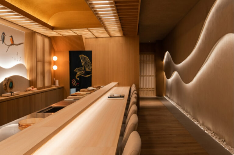 Interior of a modern Japanese omakase restaurant featuring a light wood counter, warm ambient lighting, and a wavy backlit wall design.