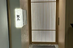A high-angle, vertical shot of the minimalist entrance to "Sushi Muni." The entryway features a traditional Japanese light-wood slatted sliding door (shoji style) set against soft, textured beige walls. To the left, a vertical rectangular lightbox made of textured washi paper glows with a warm light, displaying the Japanese kanji "無二" above the word "MUNI" in small English letters. The floor at the base of the door is lined with smooth white river stones surrounding a dark grey rectangular floor mat.