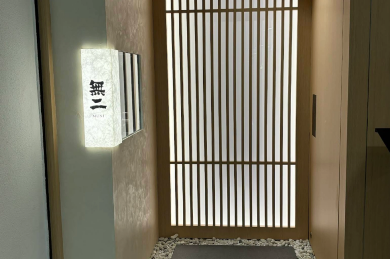 A high-angle, vertical shot of the minimalist entrance to "Sushi Muni." The entryway features a traditional Japanese light-wood slatted sliding door (shoji style) set against soft, textured beige walls. To the left, a vertical rectangular lightbox made of textured washi paper glows with a warm light, displaying the Japanese kanji "無二" above the word "MUNI" in small English letters. The floor at the base of the door is lined with smooth white river stones surrounding a dark grey rectangular floor mat.