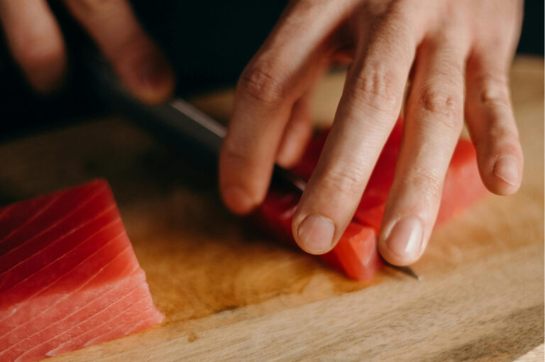 A close-up of a chef’s hands using a long, thin knife to precisely slice a block of fresh, raw red tuna on a wooden cutting board.