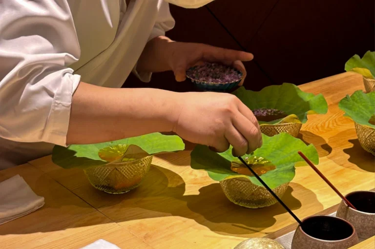 This close-up image shows a chef’s hands meticulously assembling a dish on a light-colored wooden counter. The chef, wearing a white uniform, uses a thin black tool to precisely place a garnish onto a translucent, golden-colored jelly resting on a large, vibrant green lotus leaf. The leaf is cradled in a textured, golden glass bowl, and several other identical bowls are lined up on the counter, suggesting a sequence of courses being prepared for multiple guests. In the background, the chef holds a small blue bowl filled with purple flower petals, adding a touch of natural elegance to the high-end culinary presentation.