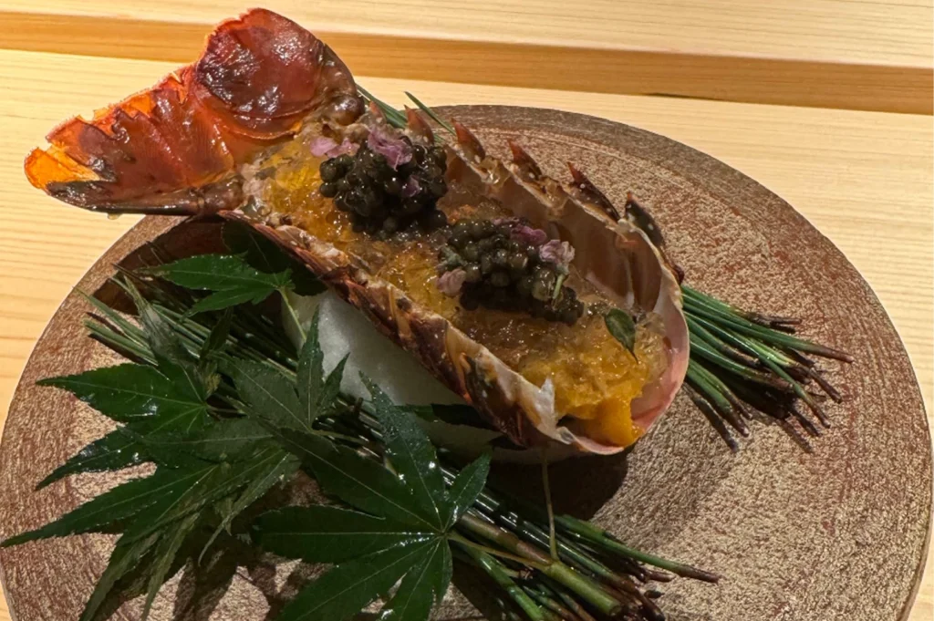 A sophisticated, artistic dish served at Hamamoto. A lobster tail shell, filled with a delicate seafood preparation topped with a generous spoonful of black caviar and tiny purple flowers, rests on a bed of ice. The plate is garnished with dark green maple leaves and pine needles, presented on a textured, golden-brown circular platter.