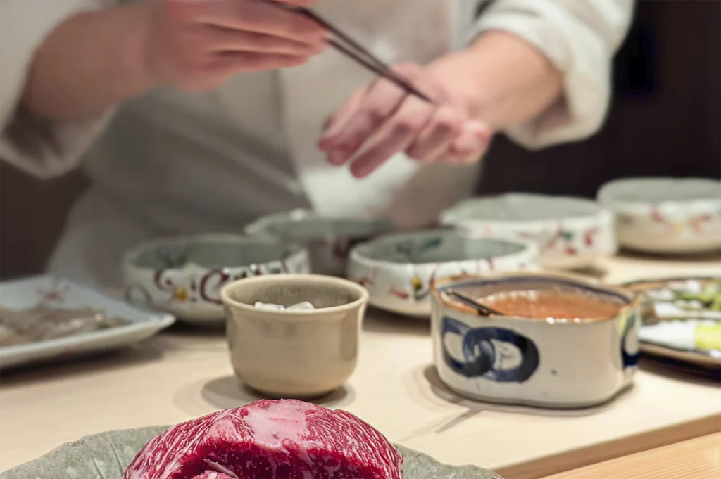 A close-up, shallow depth-of-field shot of a chef’s hands using long metal chopsticks to delicately plate food. In the foreground, a highly marbled slab of raw Wagyu beef sits on a plate. Several small, hand-painted ceramic bowls are lined up on the counter, ready for the next course of the Omakase.