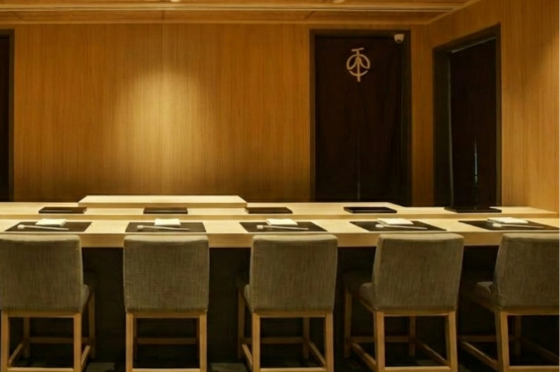 A minimalist Japanese sushi counter with light wood paneling, five place settings, and a dark curtain in the background.