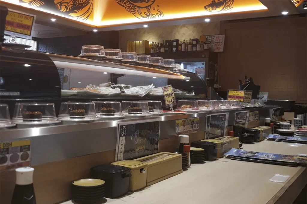 A view inside a conveyor belt sushi restaurant. The scene features a stainless steel belt system holding various small, covered plates of food. In the background, there is a lit glass display case containing fresh fish, and signs with text like "unagi" and warnings not to place sushi covers back on the belt. The counter is lined with various condiments and table settings.