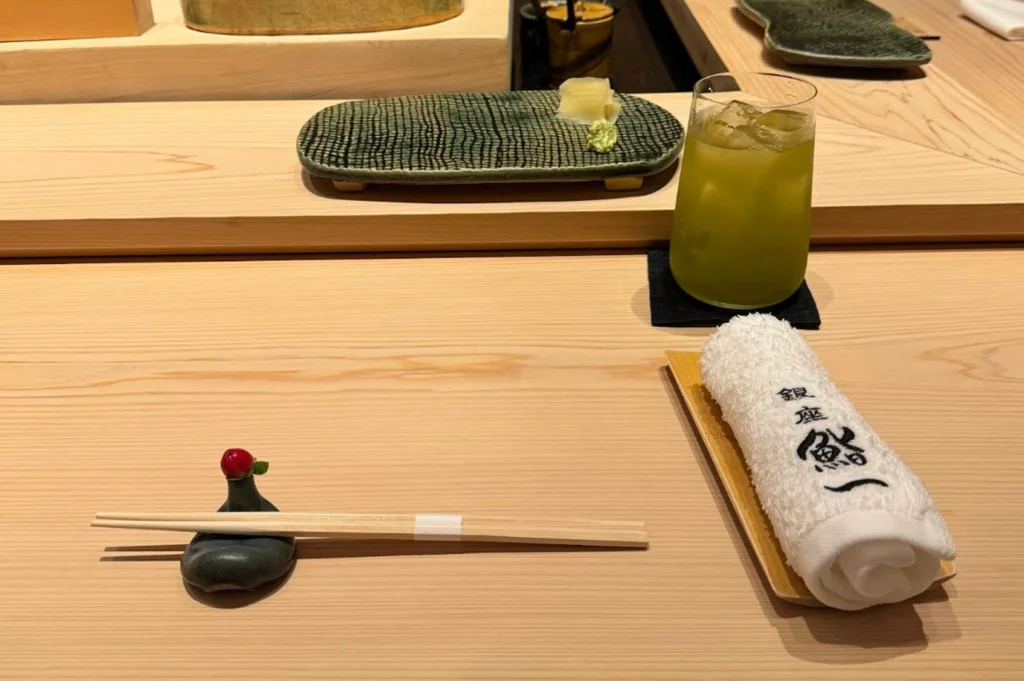 A high-angle, close-up shot captures a minimalist and elegant place setting at a traditional Japanese omakase sushi counter made of light-colored, smooth-grained wood. In the foreground, a pair of light wood chopsticks rests horizontally on a dark, pebble-shaped ceramic holder adorned with a single small red berry. To the right, a white, rolled-up oshibori (hot towel) with black Japanese calligraphy sits on a thin wooden tray. Beside it, a frosted glass filled with vibrant iced green tea rests on a dark square coaster. In the mid-ground, a textured, dark green ceramic rectangular plate holds small mounds of pickled ginger and wasabi, positioned on a raised wooden tier of the counter. The background is softly blurred, showing more of the clean wooden counter and a glimpse of ceramic dishes, creating a serene and sophisticated dining atmosphere.