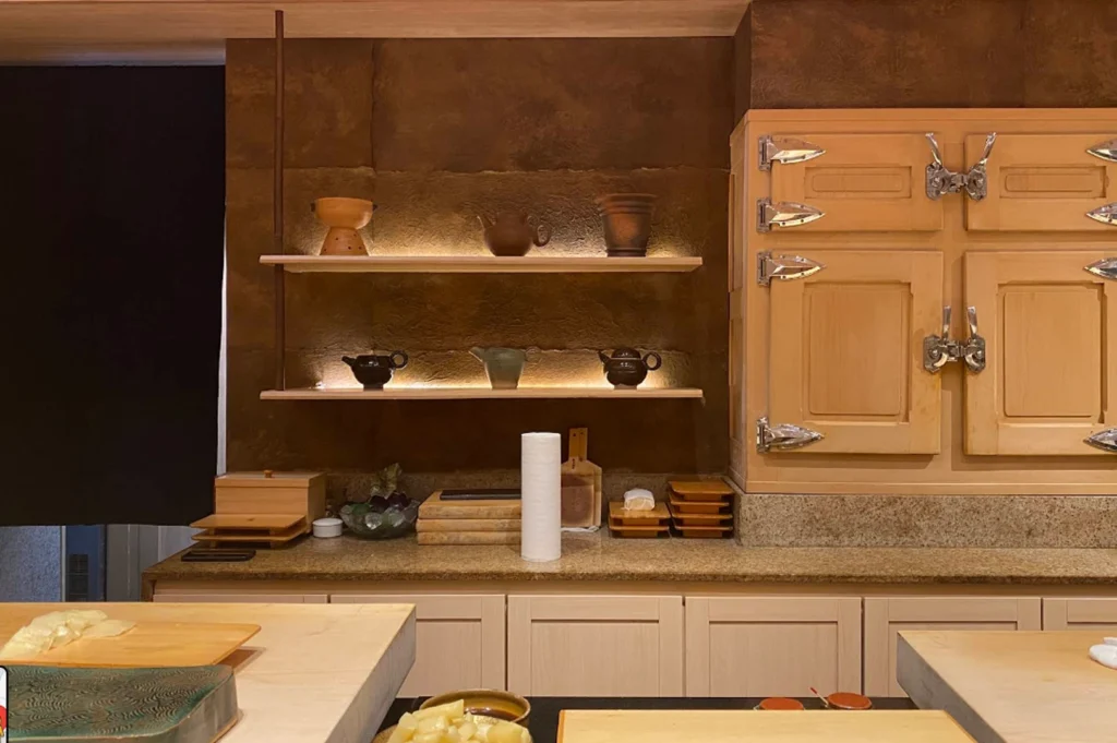 An interior view of the professional sushi preparation area. The background features a dark, earthy-textured wall with two floating wooden shelves holding artisanal ceramic teapots and bowls, illuminated by warm under-shelf lighting. To the right is a large, traditional wooden ice chest (refrigerator) with heavy metallic latches. In the foreground, clean light-wood counter spaces are set up with essential tools, including a roll of paper towels, wooden trays, and small containers of ginger.