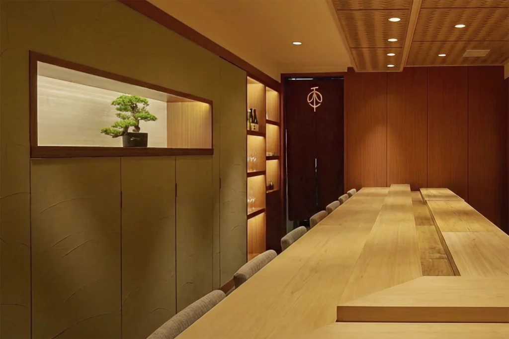 An elevated view of a long, pristine light-wood sushi counter lined with grey upholstered chairs. The room is bathed in warm, golden light, featuring a recessed wall niche holding a small bonsai tree and a lit shelving unit stocked with glassware and sake bottles in the background.