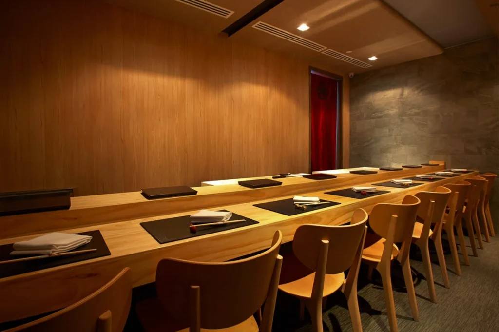 A long, perspective shot of an empty, upscale sushi bar featuring a continuous light wood counter that stretches toward a red curtain at the far end. Each seat is prepared with a black placemat, a folded white napkin, and chopsticks resting on a small holder. The room is illuminated by warm, recessed ceiling lights that highlight the smooth wood grain of the walls and the modern, light wood chairs tucked neatly under the counter.