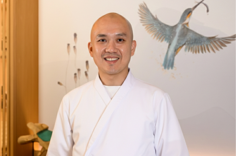 A smiling male sushi chef in a white traditional uniform standing in front of a wall decorated with a kingfisher illustration.
