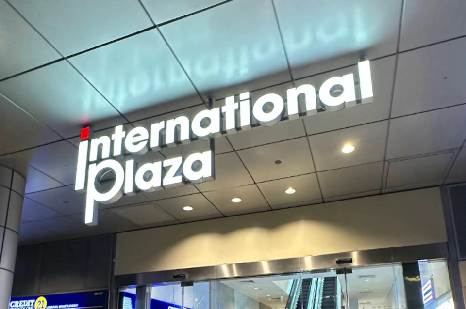 A low-angle shot of the glowing exterior signage for "International Plaza." The words "International Plaza" are rendered in large, white 3D block letters with a small red square dotting the "i." The sign is mounted against a grid-patterned ceiling with recessed warm spotlights. Its reflection is visible on the polished ceiling tiles above. Below the sign, glass automatic doors reveal a glimpse of an indoor escalator and the bright interior of a commercial building.