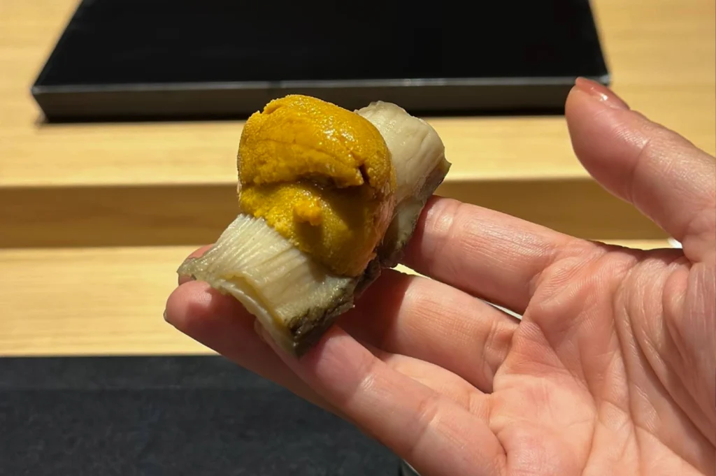A person’s hand holds a single, exquisite piece of sushi-style seafood against a soft-focus background of a wooden sushi counter. The bite consists of a thick, pale slice of steamed or braised abalone topped with a generous, vibrant orange mound of fresh uni (sea urchin). The textures are prominently displayed, from the smooth, slightly ridged surface of the abalone to the creamy, delicate lobes of the uni. The lighting is warm and direct, highlighting the freshness and luxury of the ingredients.