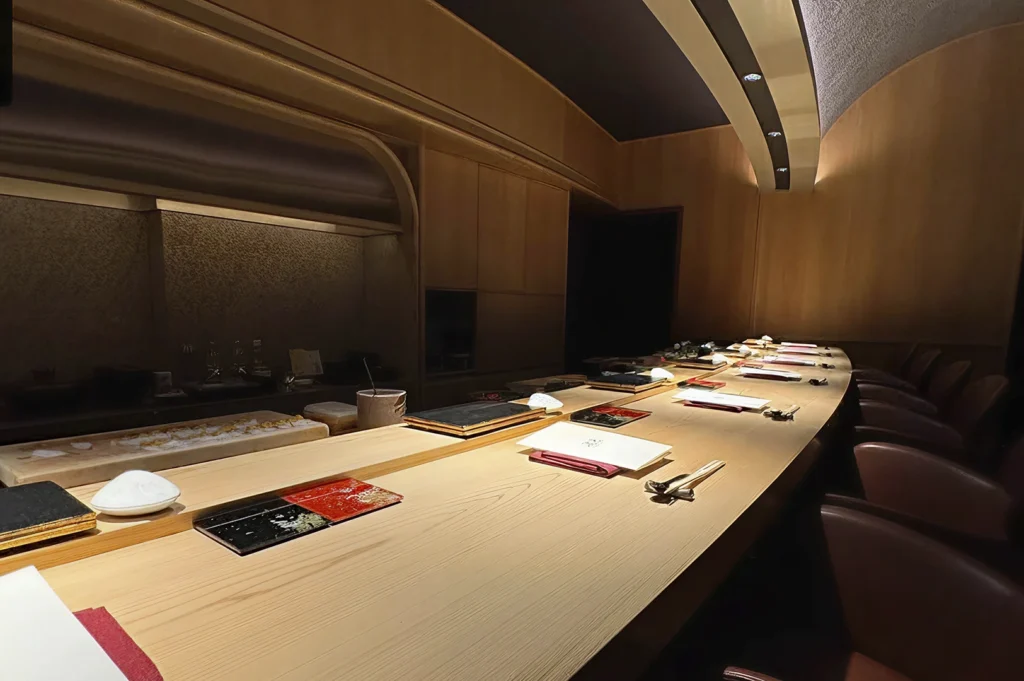 A wide-angle view of the dining room at Hamamoto. The space is warm and intimate, featuring a smooth, curved wooden counter that follows the contour of the room. Soft, recessed lighting highlights the wood grain and the minimalist place settings, creating a serene, upscale atmosphere.