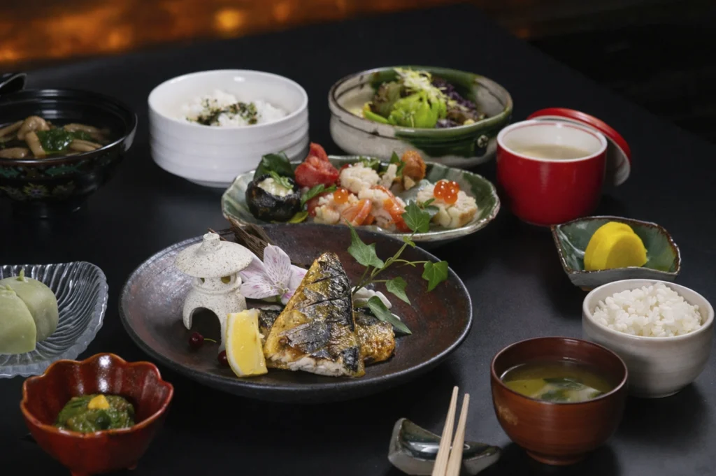 A top-down view of a traditional Japanese meal spread on a dark table. The centerpiece is a ceramic plate holding grilled mackerel garnished with a lemon wedge and a decorative miniature stone lantern. Surrounding it are small bowls containing miso soup, white rice, pickled vegetables, and a vibrant seafood salad topped with ikura (salmon roe).