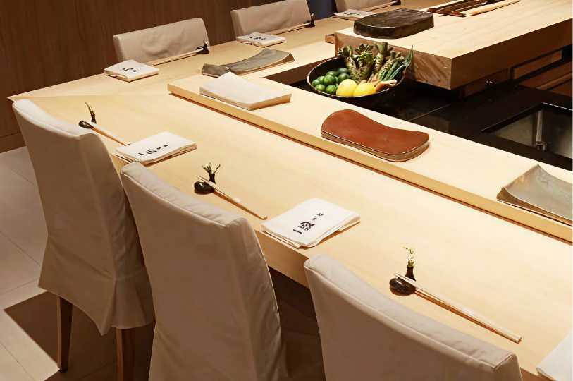 A close-up, angled view of a clean wooden omakase counter featuring white napkins, chopsticks on rests, and a bowl of fresh wasabi root and citrus.
