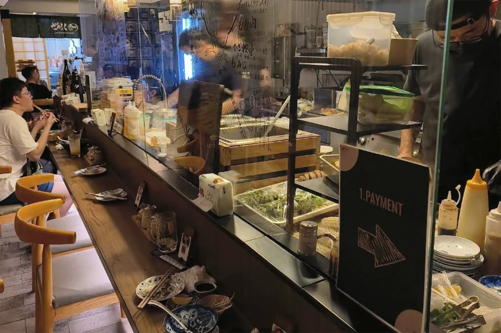 A perspective from inside a Japanese restaurant, looking across a long wooden bar counter toward an open kitchen area behind a glass partition. A customer sits at the bar on the left, eating. Behind the glass, a chef is working, and there is a sign that reads "1. PAYMENT" with a downward-pointing arrow.