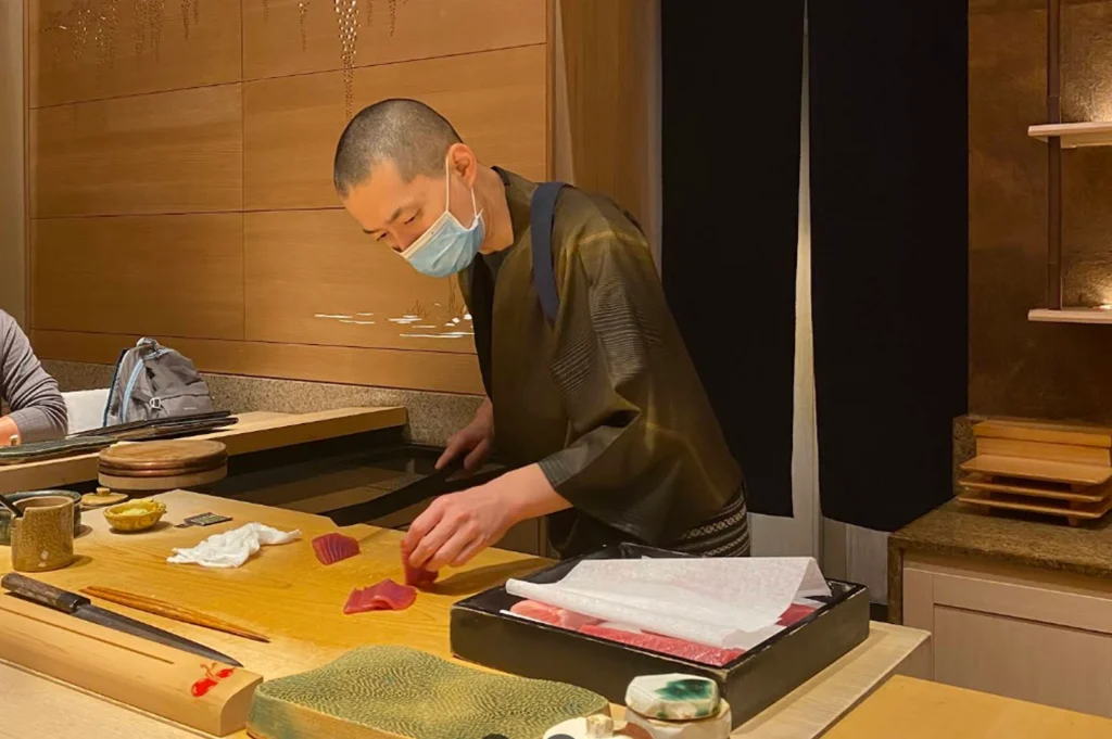 A sushi chef meticulously at work behind a bright wooden counter. The chef, with a shaved head and wearing a blue surgical mask and a dark traditional Japanese work tunic (samue), leans over a large wooden cutting board. He is precisely slicing bright red pieces of tuna (maguro) with a long sushi knife. In front of him is a black lacquer box containing several cuts of fresh, marbled fish protected by paper.