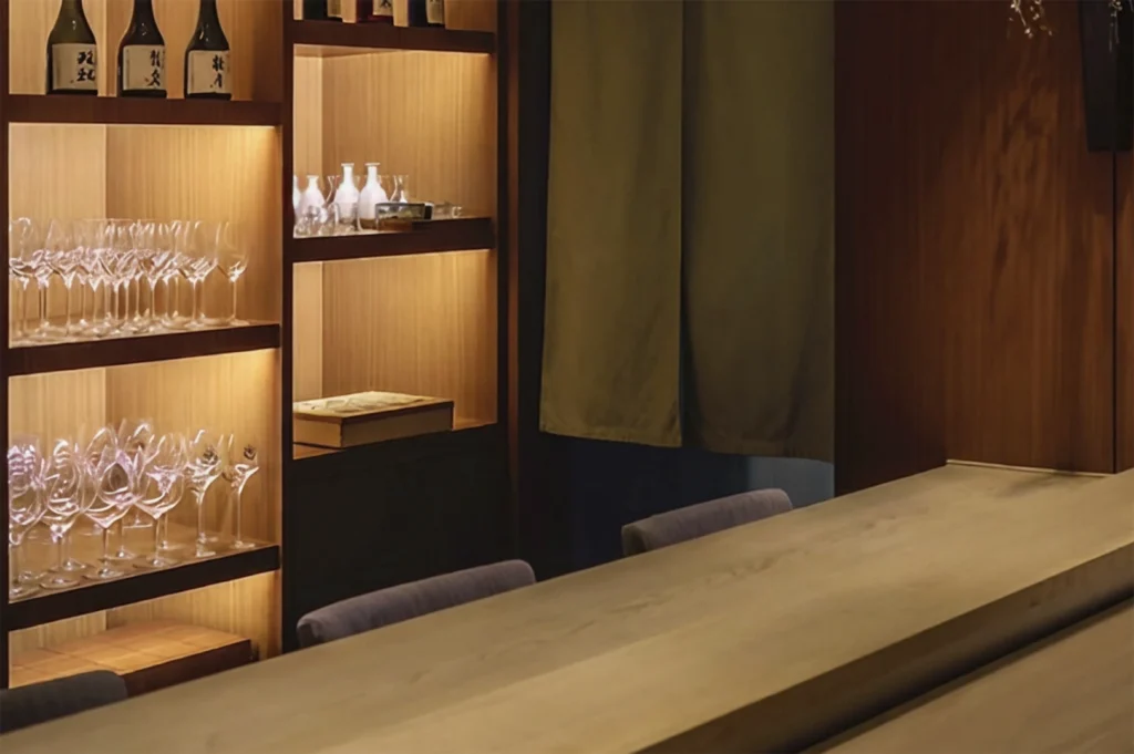 A close-up of a high-end sushi bar area showing a smooth, light-colored wooden counter in the foreground. In the background, illuminated wooden shelves display rows of elegant wine glasses, ceramic sake carafes, and premium sake bottles, partially framed by a dark green hanging fabric divider.