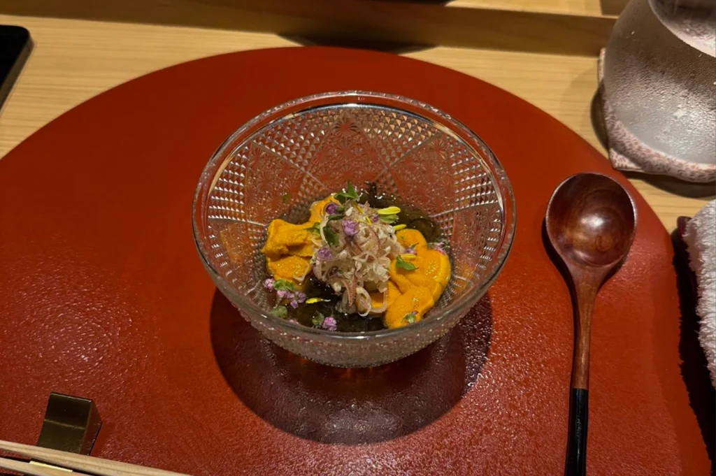A top-down, close-up shot of an intricate Japanese dish served in a decorative, clear cut-glass bowl placed on a textured reddish-brown charger. Inside the bowl, bright orange lobes of fresh uni (sea urchin) are nestled atop a dark, jelly-like base, garnished with delicate purple flowers and finely shredded greens. A long-handled wooden spoon rests to the right of the bowl, and the corner of a wooden table and a white towel are visible at the edges of the frame.