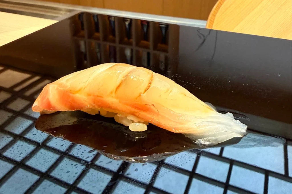 This close-up, high-angle shot features a single piece of nigiri sushi meticulously placed on a glossy, reflective black lacquer serving board. The sushi consists of a translucent, pale orange and white slice of fish—gently scored and brushed with a light glaze of soy sauce—draped over a small, compact bed of seasoned white rice. The polished black surface of the board creates a sharp reflection of the sushi piece, while the grid pattern of the restaurant’s illuminated shoji ceiling is clearly mirrored across the bottom left of the platform. In the soft-focused background, the warm tones of a wooden chair and the gridded sake bottle display are subtly visible, keeping the sharp focus entirely on the delicate textures of the fish.