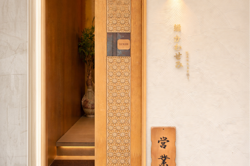 A close-up of a wooden sliding door with intricate geometric carvings and vertical Japanese calligraphy on the adjacent textured white wall.