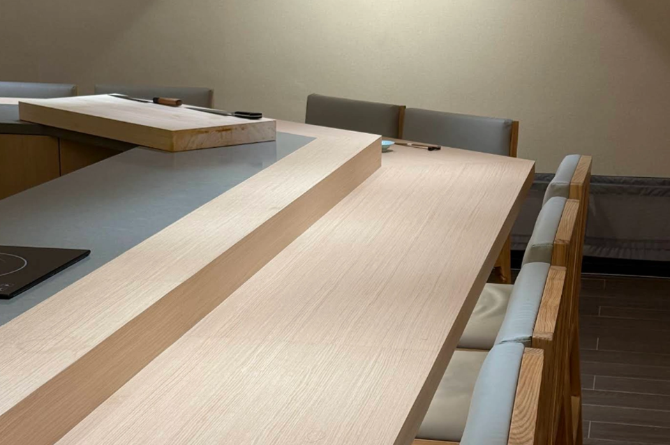 An interior shot of an empty, upscale sushi counter. The L-shaped counter is made of smooth, light-toned wood with a clean, minimalist design. On the raised preparation section in the background, a thick wooden cutting board holds two long, professional sushi knives (yanagiba). The seating area features light grey upholstered chairs with wooden frames. The background is a plain, neutral-colored wall, and the floor is made of dark wood-style planks, creating a serene and focused dining atmosphere.