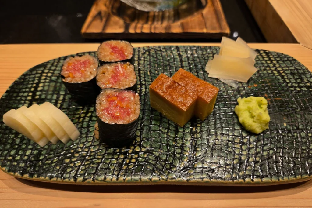 This overhead shot captures an assortment of Japanese delicacies presented on a rectangular, textured green and black ceramic plate. The plate holds four pieces of maki (sushi rolls) filled with bright pink tuna and seasoned rice, two thick slices of golden-brown tamagoyaki (sweet omelet) that look almost like cake, a neat pile of translucent pickled ginger, a small mound of bright green wasabi, and several slices of pale, pickled radish. The arrangement is clean and artistic, set against the backdrop of a light wood counter that is typical of a traditional omakase setting.