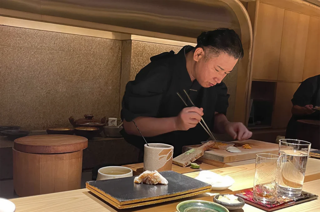 Chef Kazuhiro Hamamoto standing behind the wooden counter in his kitchen. Dressed in a black uniform, he is focused intently on preparing a piece of nigiri sushi using long, thin chopsticks. The workstation is neat, featuring a wooden bowls of ingredients, and a partially plated dish on a dark slate board.