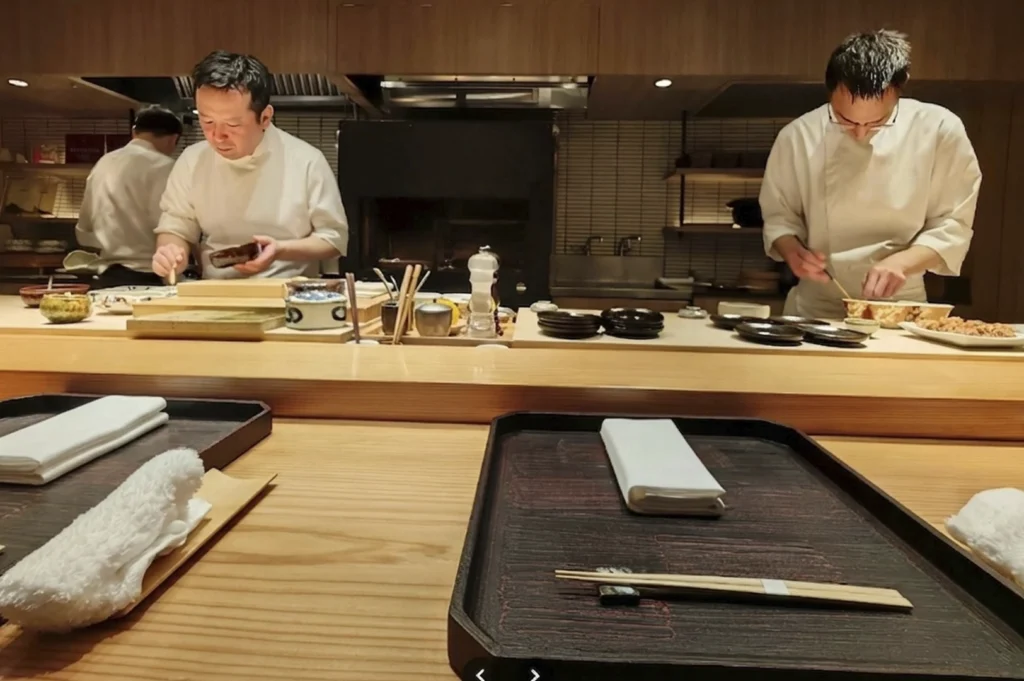 Two chefs in traditional white uniforms work behind a clean, light-wood Kappo counter. One chef is focused on plating a small dish while the other prepares ingredients. In the foreground, the customer's perspective shows a dark wooden tray with a neatly folded white napkin, a damp hand towel (oshibori), and chopsticks on a rest.