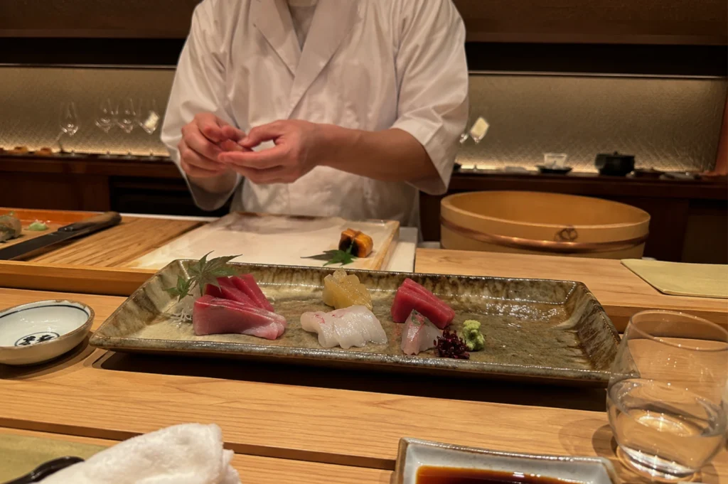 A close-up shot captures an upscale omakase dining experience where a sushi chef wearing a crisp white jacket stands behind a smooth, light-colored wooden counter, meticulously shaping food with his hands. In the immediate foreground, a long, rustic ceramic platter displays an elegant assortment of sashimi, including rich red and pink tuna slices, translucent white fish, a dollop of green wasabi, and a small decorative leaf. The counter also holds a small dipping dish with Japanese kanji, a glass of water, a saucer of dark soy sauce, and a rolled white hand towel. In the background, a traditional large wooden sushi rice tub (hangiri) and clean preparation surfaces sit under warm, soft lighting, highlighting the intimate and artistic nature of Japanese fine dining.