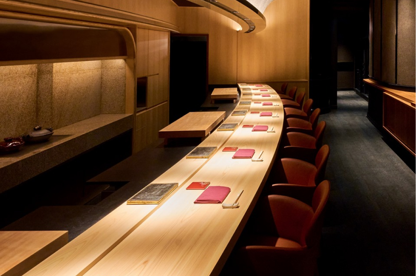 A long, curved wooden sushi bar illuminated by warm overhead lighting, with elegant red chairs and formal place settings.