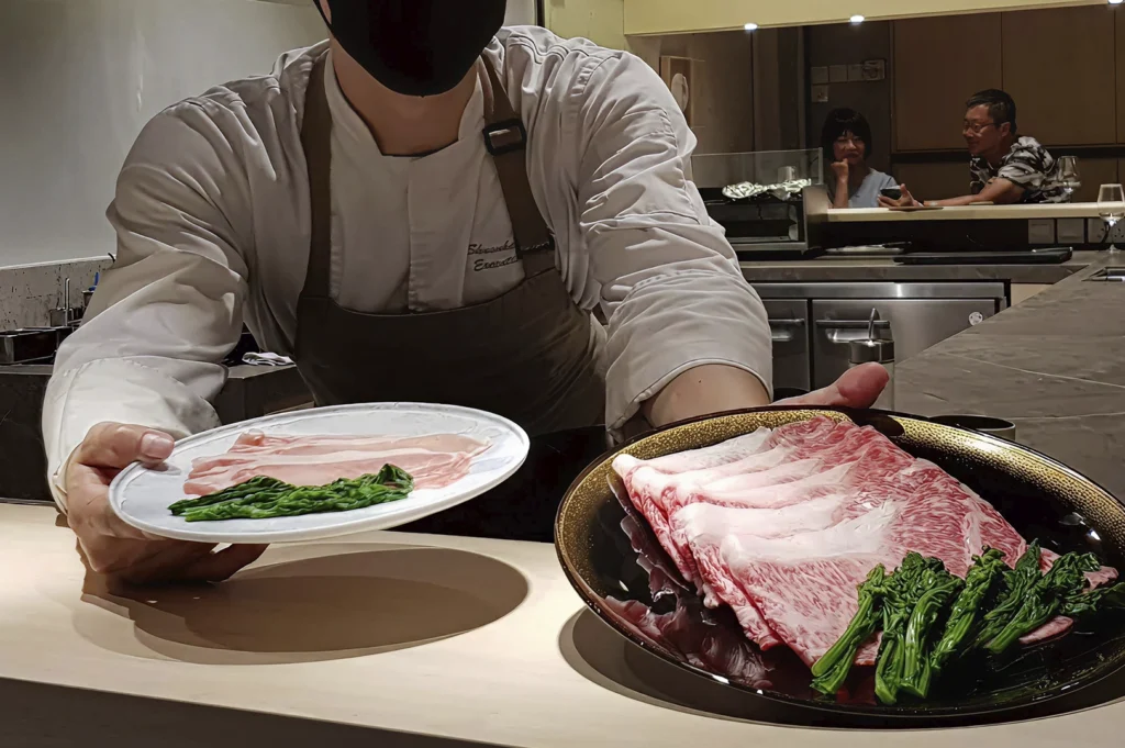 A close-up shot of a sushi chef, wearing a black face mask and a white uniform with an apron, holding out two plates of food for a customer. The plate in his left hand holds thin slices of raw meat with a small portion of green vegetables, while the plate in his right hand features a larger portion of beautifully marbled wagyu beef arranged with cooked greens.