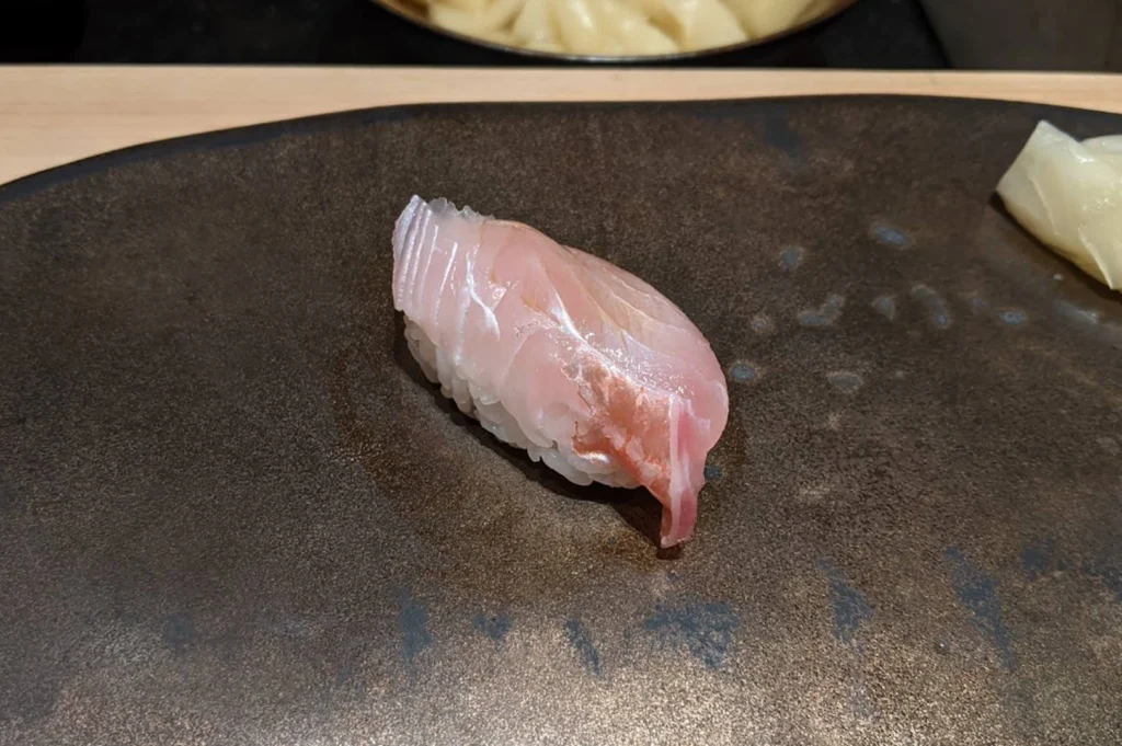 A single piece of Kasugo (young sea bream) nigiri. The fish is translucent with a soft, iridescent pink and silver skin, delicately scored to reveal the white flesh beneath. It rests on a small mound of seasoned sushi rice. The sushi is centered on a rustic, dark brown stoneware plate with a metallic, textured finish. In the background, a small portion of thinly sliced pickled ginger sits on the edge of the plate, and a wooden counter is visible at the top. The lighting is soft and natural, emphasizing the moist texture of the fish and the individual grains of rice.