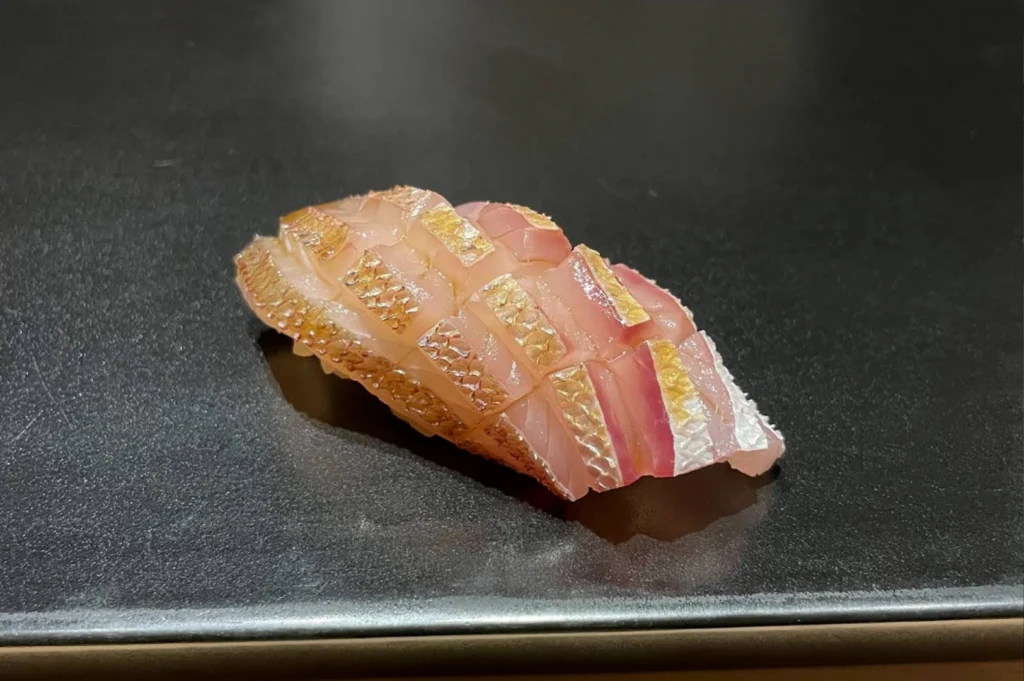 A macro, close-up shot of a single piece of nigiri sushi resting on a sleek, matte black ceramic surface. The fish, likely a type of snapper or sea bream, features translucent pinkish-white flesh with a beautifully scored skin that has been lightly seared to a golden-brown hue. The intricate texture of the fish and the delicate glisten of a glaze are highlighted against the dark, minimalist background, emphasizing the precision of the preparation.