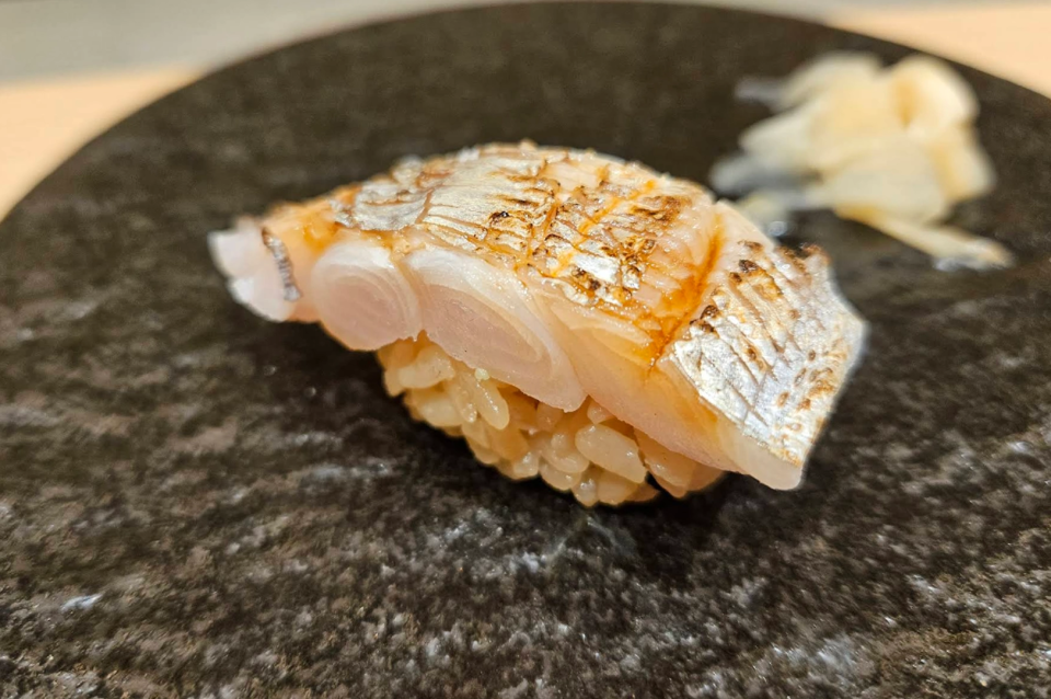 A macro, eye-level close-up of a single piece of nigiri sushi served on a textured black ceramic plate. The fish, likely a type of white fish or silver-skinned fish like shime saba or kamasu, is expertly scored with delicate cross-hatch cuts and lightly seared on top to reveal a glistening, golden-brown finish. It sits atop a small hand-pressed mound of seasoned sushi rice. In the soft-focus background, a small pile of pale pickled ginger (gari) is visible.