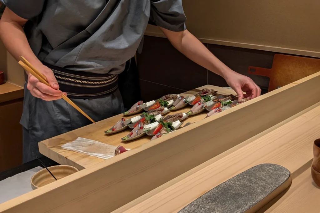 A chef in a slate-blue traditional uniform and a decorative dark belt is shown from the waist down, working behind a clean, light wood sushi counter. He is using long wooden chopsticks and his fingers to carefully arrange a series of small, silver, spoon-like serving vessels. Each vessel contains a delicate slice of sashimi topped with a sliver of tomato, fresh greens, and tiny red seeds, likely pomegranate. The repetitive, orderly arrangement of the small plates emphasizes the precision and repetitive craftsmanship involved in a professional omakase service.