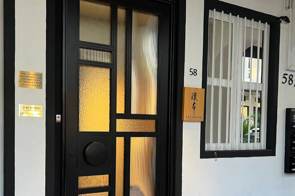 The exterior entrance to Hamamoto restaurant. A sleek, modern black door with textured glass panels sits next to a white-painted wall with a vertical white-barred window. A wooden sign with the name "Hamamoto" in English and Japanese kanji hangs on the wall beside the door, with the number "58" displayed clearly above it.