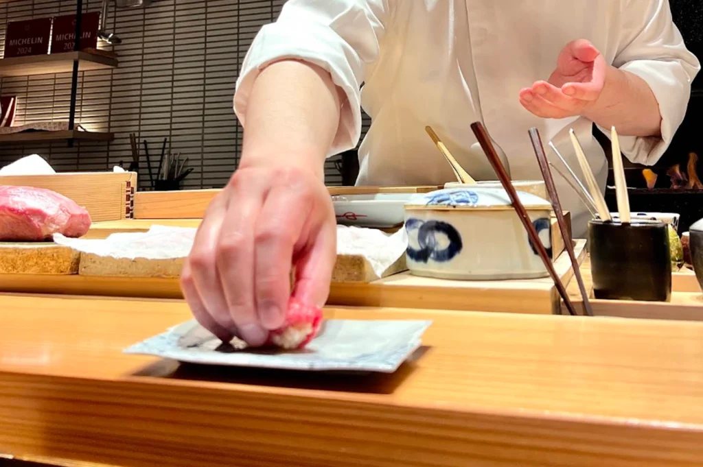 A chef's hand is captured in motion, placing a fresh piece of nigiri sushi onto a small, rectangular ceramic plate. The background shows various kitchen tools, including wooden ingredient boxes and ceramic containers, under warm lighting. A Michelin 2024 plaque is visible on the back wall.