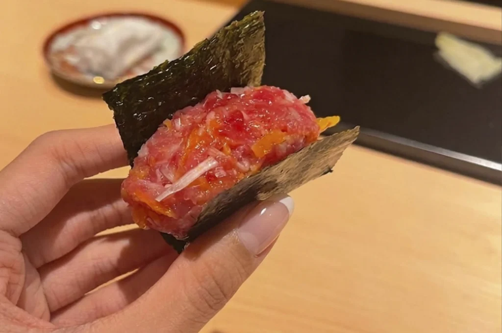 A close-up photograph of a person's hand holding a crisp, dark green nori (seaweed) handroll filled with a generous amount of fatty tuna tartare (negitoro). The tuna is vibrant pink and finely chopped, mixed with small orange flecks, possibly pickled radish. The hand has a clean, minimalist manicure with white tips. The background is a soft-focus view of a light wooden table and a small ceramic dish, keeping the focus entirely on the texture of the fresh seafood and the seaweed.