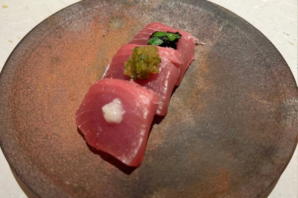 A top-down, slightly angled shot of three thick slices of fresh, pink sashimi—likely medium-fatty tuna (chu-toro)—arranged in a row on a rustic, dark-textured ceramic plate. Each slice is topped with a different delicate garnish: a small dollop of white paste (possibly ginger or onion), a bright green herb relish, and a dark, glossy topping with a small green leaf. The plate has an earthy, metallic patina, contrasting with the clean, white speckled tabletop beneath it.