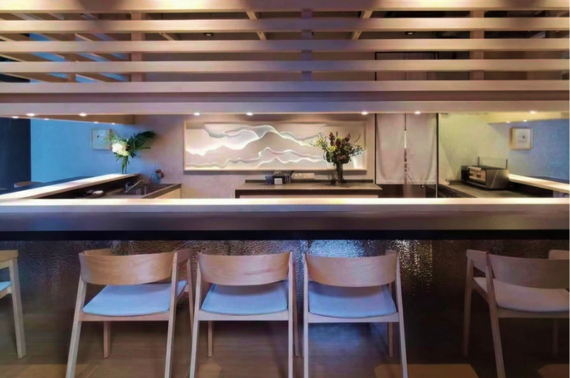 A modern sushi counter viewed through a wooden slatted partition, featuring a textured silver base and a backlit mountain-style wall art.