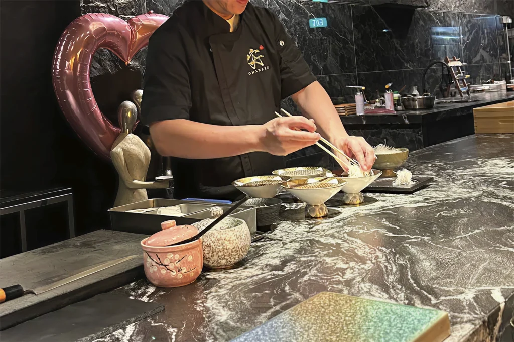 A chef in a black uniform with "OMAKASE" embroidered on the chest stands behind a polished, dark-veined stone counter. He is using chopsticks to carefully place delicate, thin noodles into small, elegant serving bowls. To his left, there is a large, metallic, heart-shaped balloon, and his workspace is clean and organized with various prep bowls.