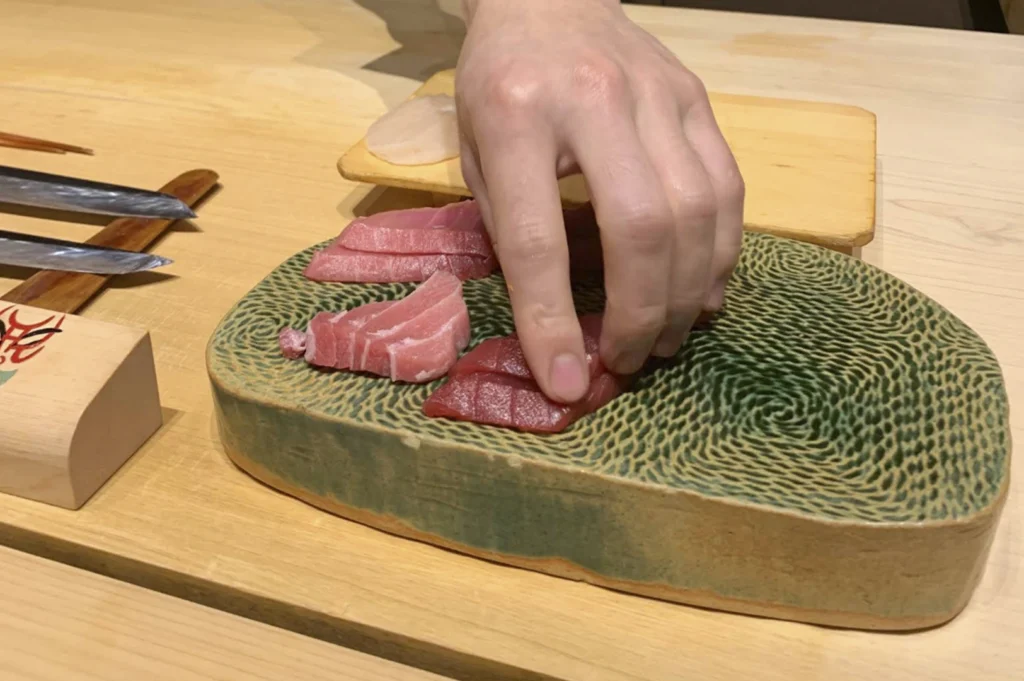 A close-up action shot focusing on the chef's hand as he arranges slices of fresh sashimi. The fish, appearing to be different fatty grades of tuna (Otoro and Chutoro), is placed on a unique, elevated ceramic serving block with a striking green-and-tan swirling, textured pattern. Two long, slender sushi knives with dark handles rest on a wooden block to the left, highlighting the precision of the craft.
