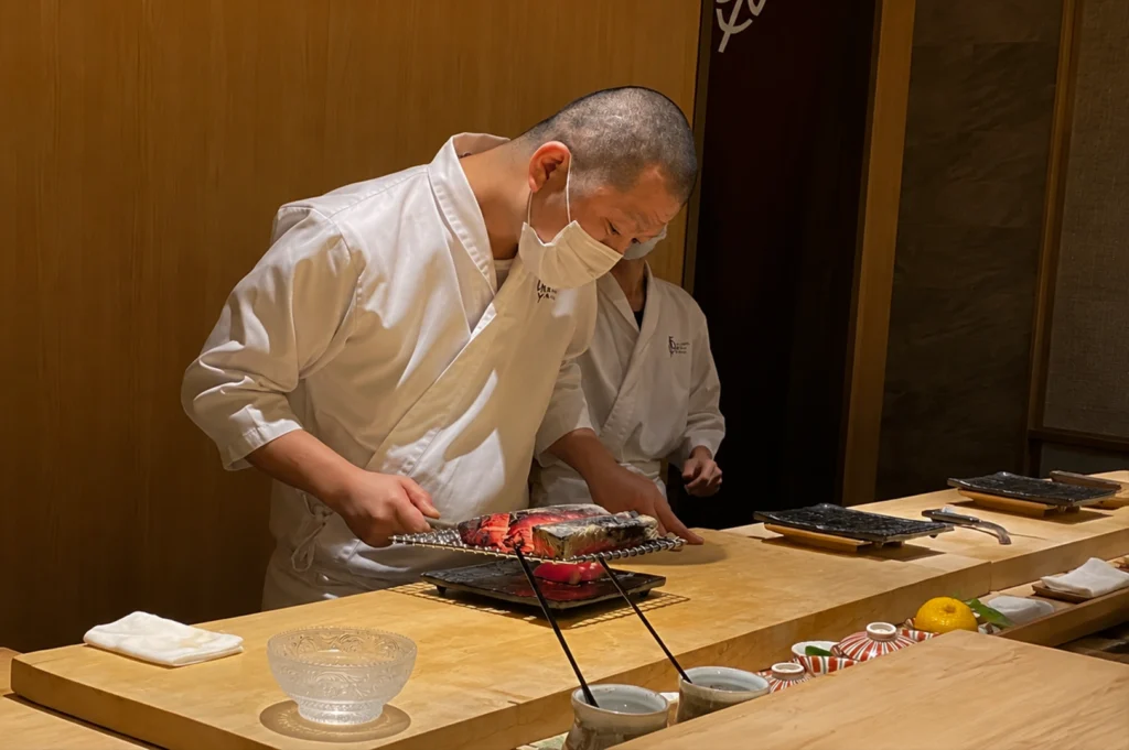 A long, perspective shot of an empty, upscale sushi bar featuring a continuous light wood counter that stretches toward a red curtain at the far end. Each seat is prepared with a black placemat, a folded white napkin, and chopsticks resting on a small holder. The room is illuminated by warm, recessed ceiling lights that highlight the smooth wood grain of the walls and the modern, light wood chairs tucked neatly under the counter.