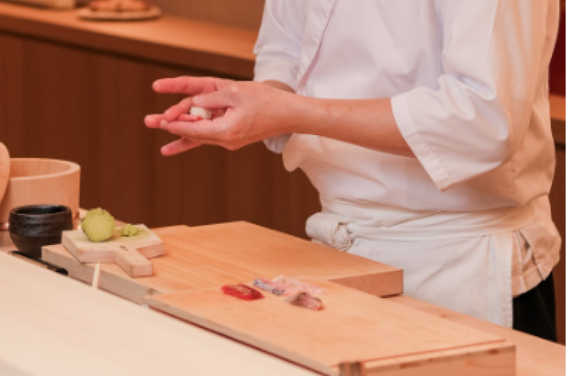 A close-up of a sushi chef's hands delicately shaping nigiri over a wooden preparation board with fresh fish slices and wasabi.